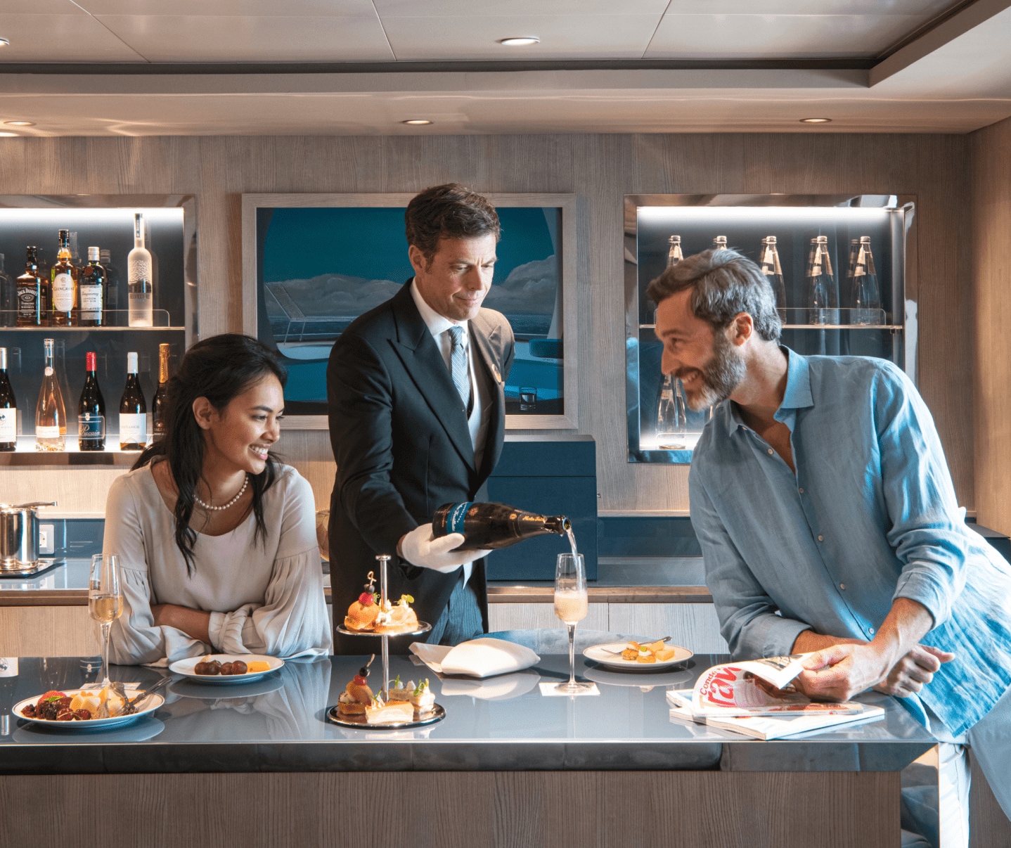 A MSC Yacht Club Butler in a distinguished black suit and white gloves serves tea with finger foods to a couple in a private yacht club area. They relax on a couch, savoring the moment while enjoying breathtaking ocean views.